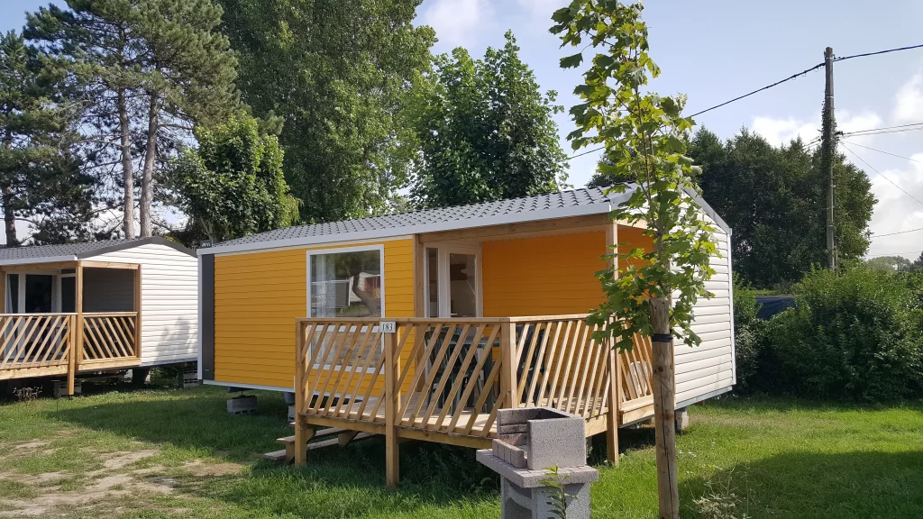 Mobil-home jaune avec terrasse et arbres environnants.