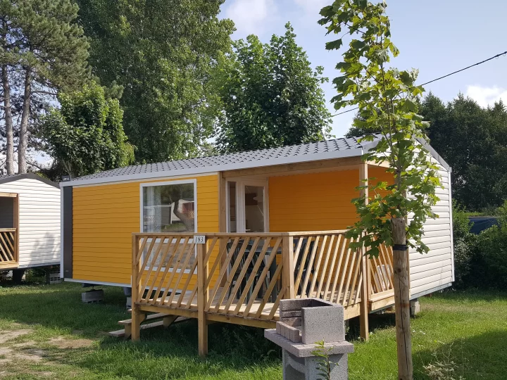 Mobil-home jaune avec terrasse et arbres environnants.