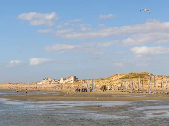 Plage animée avec voiliers et dunes en arrière-plan.